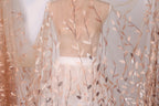 a mannequin wearing a white dress with a sheer, leaf-like fabric draped over it. The fabric has a delicate, floral pattern in shades of peach and white.