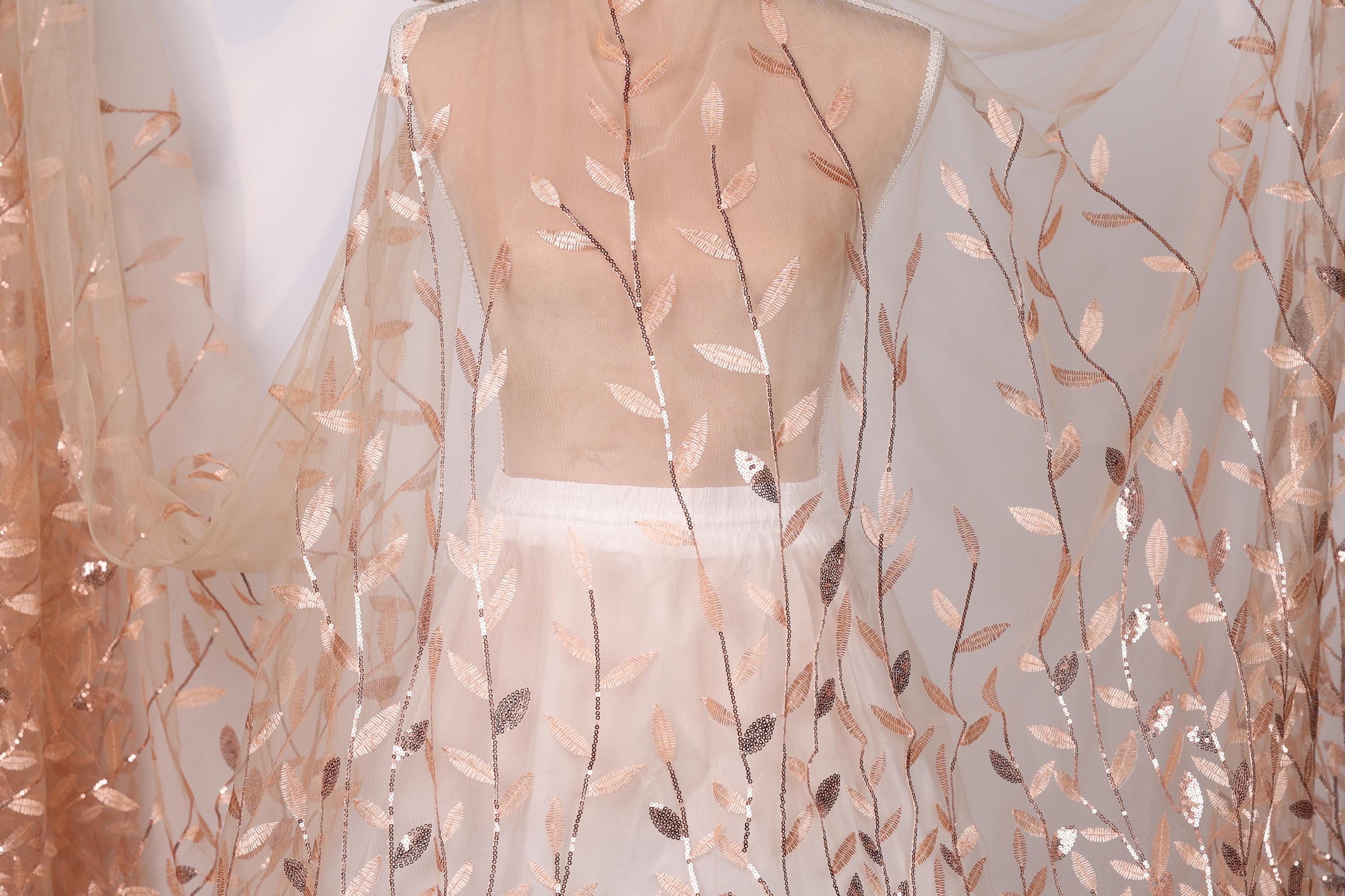a mannequin wearing a white dress with a sheer, leaf-like fabric draped over it. The fabric has a delicate, floral pattern in shades of peach and white.