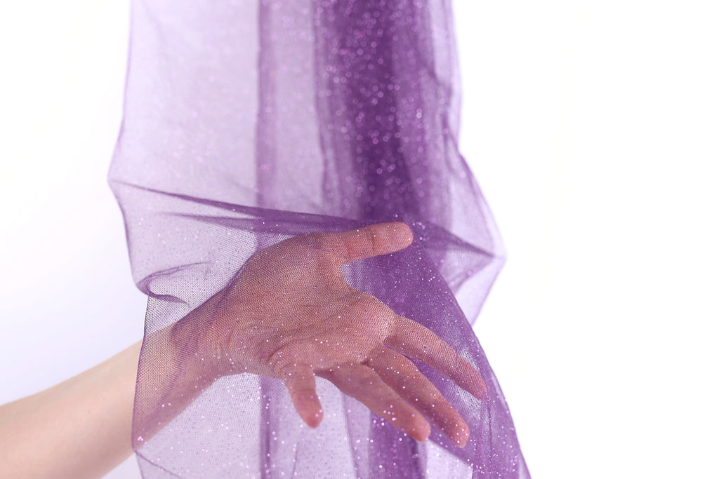 A hand is holding a purple fabric with sparkles, partially covering the hand and creating a sense of depth and texture.