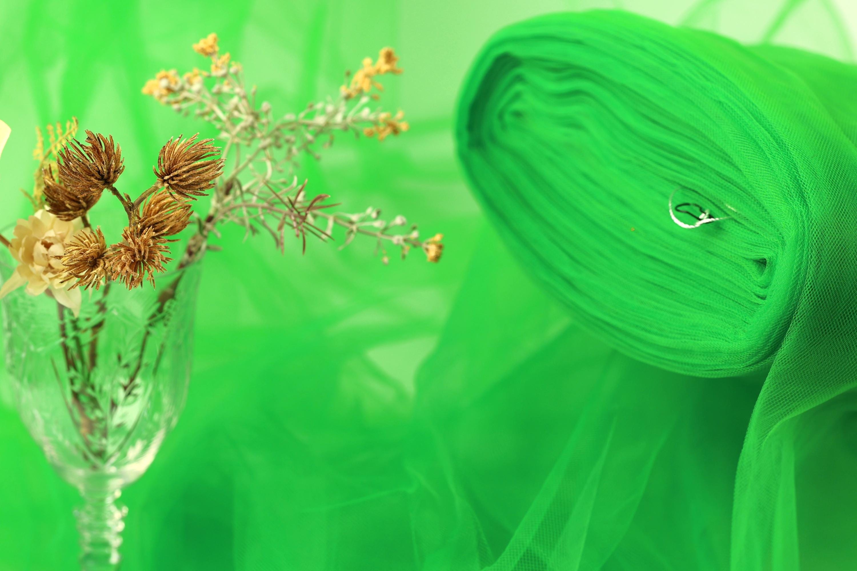 a glass vase filled with dried flowers and a green fabric or material, possibly a piece of tulle, against a green background.