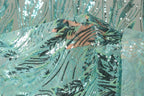 a close-up view of a fabric with a pattern of sequins and beads in shades of blue and green. The fabric appears to be a sheer material, with a floral or leaf-like design. The background is blurred, drawing focus to the fabric with its intricate details.