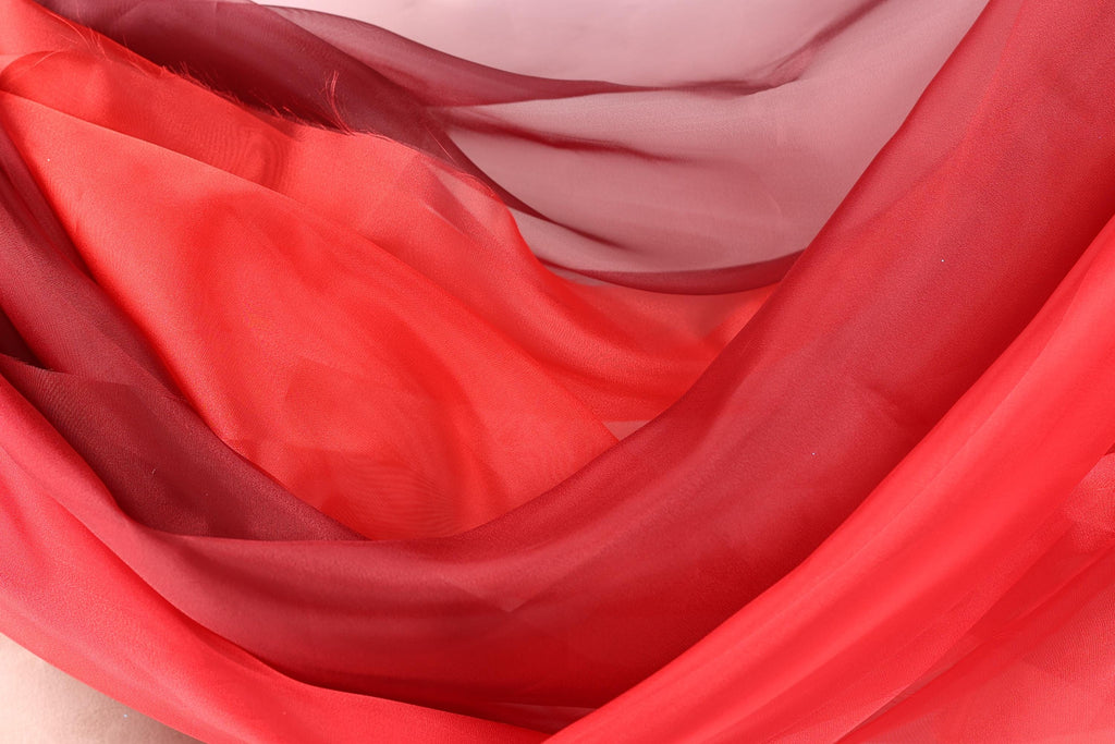 a close-up view of a red fabric, possibly silk or chiffon, with a mix of red and maroon hues. The fabric appears to be flowing and draped, creating a sense of movement and softness.