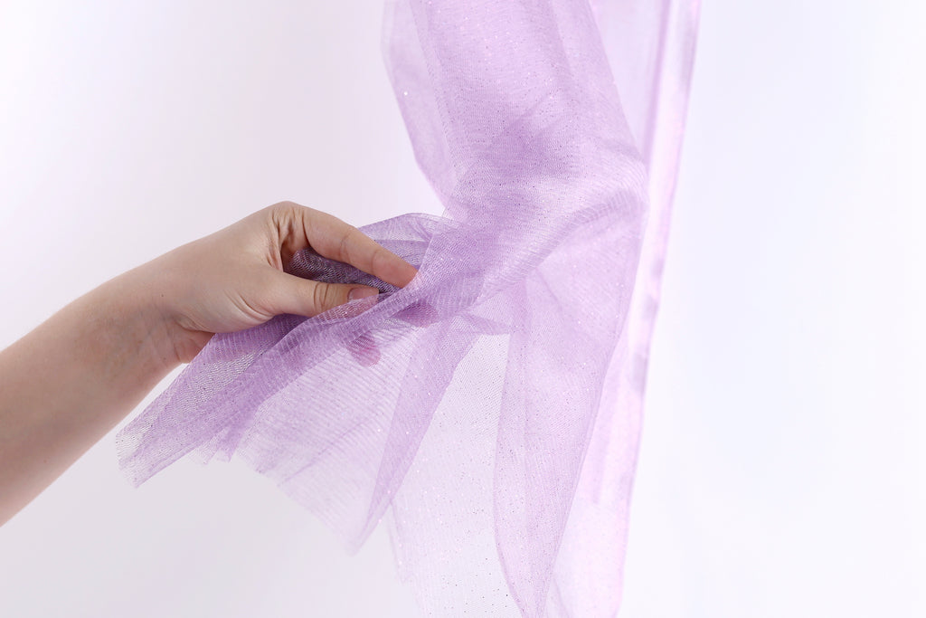 A hand is holding a piece of purple tulle fabric against a white background.