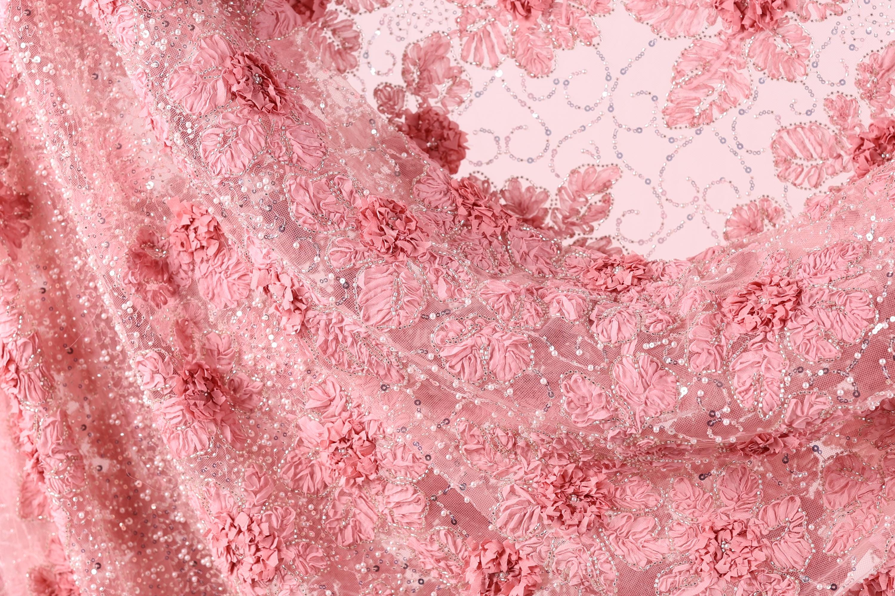 a close-up view of a delicate, pink lace fabric with a floral pattern. The fabric appears to be made of sheer material, giving it a soft and delicate appearance. The lace is intricately designed with pink flowers and leaves, creating a visually appealing and elegant design.