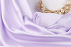 A close-up view of a soft, flowing purple satin fabric, with a rolled-up purple towel resting on top of it. In the background, there is a wreath made of dried flowers, adding a natural element to the scene.