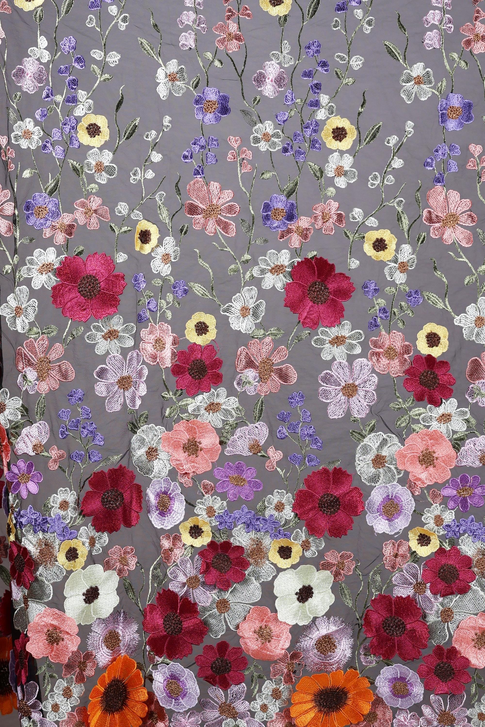 a close-up view of a fabric with a vibrant floral pattern. The fabric is adorned with a variety of colorful flowers, including red, pink, white, and yellow flowers, as well as purple and orange flowers. The flowers are intricately embroidered or sewn onto the fabric, creating a visually striking and detailed design. The image does not contain any text or other discernible objects, and the focus is solely on the floral pattern on the fabric.