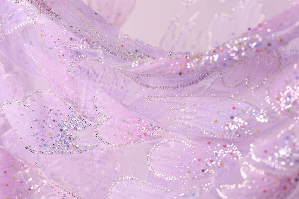 The image is a close-up of a fabric with a purple and pink color scheme, adorned with small, multicolored beads or sequins that create a sparkling, textured effect.