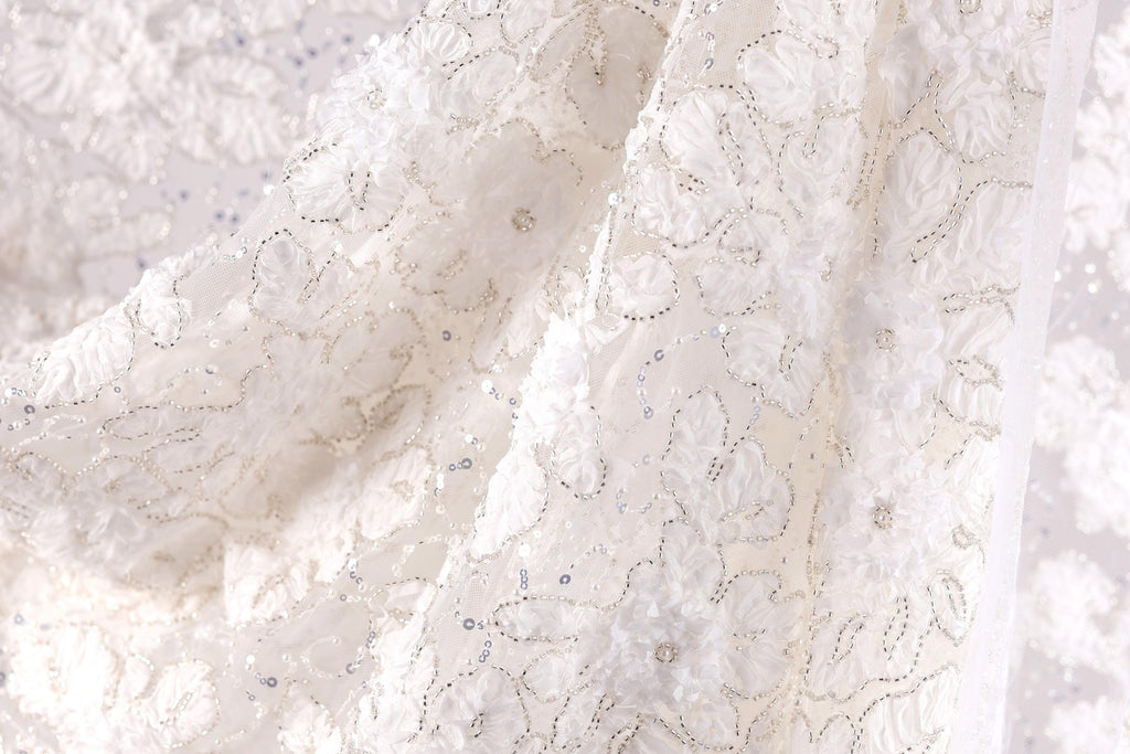 a close-up view of a delicate, white lace fabric with intricate floral patterns. The fabric appears to be made of a soft, silky material and is adorned with small, sparkling embellishments. The image does not contain any text or other discernible objects, and the focus is solely on the intricate details of the lace fabric.