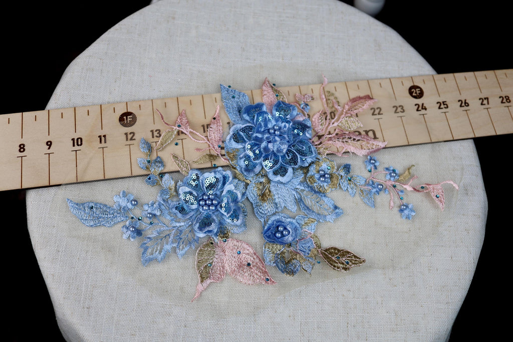 a close-up view of a delicate floral embroidery with blue and pink flowers, placed on a white fabric surface. A wooden ruler is visible in the background, likely used for measuring the size of the embroidered flowers.