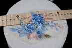 a close-up view of a delicate floral embroidery with blue and pink flowers, placed on a white fabric surface. A wooden ruler is visible in the background, likely used for measuring the size of the embroidered flowers.