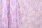 a close-up view of a fabric with a floral pattern in shades of pink and purple. The fabric appears to be made of a sheer material, with a delicate and intricate design. The pattern consists of multiple flowers and leaves, creating a visually appealing and textured appearance.