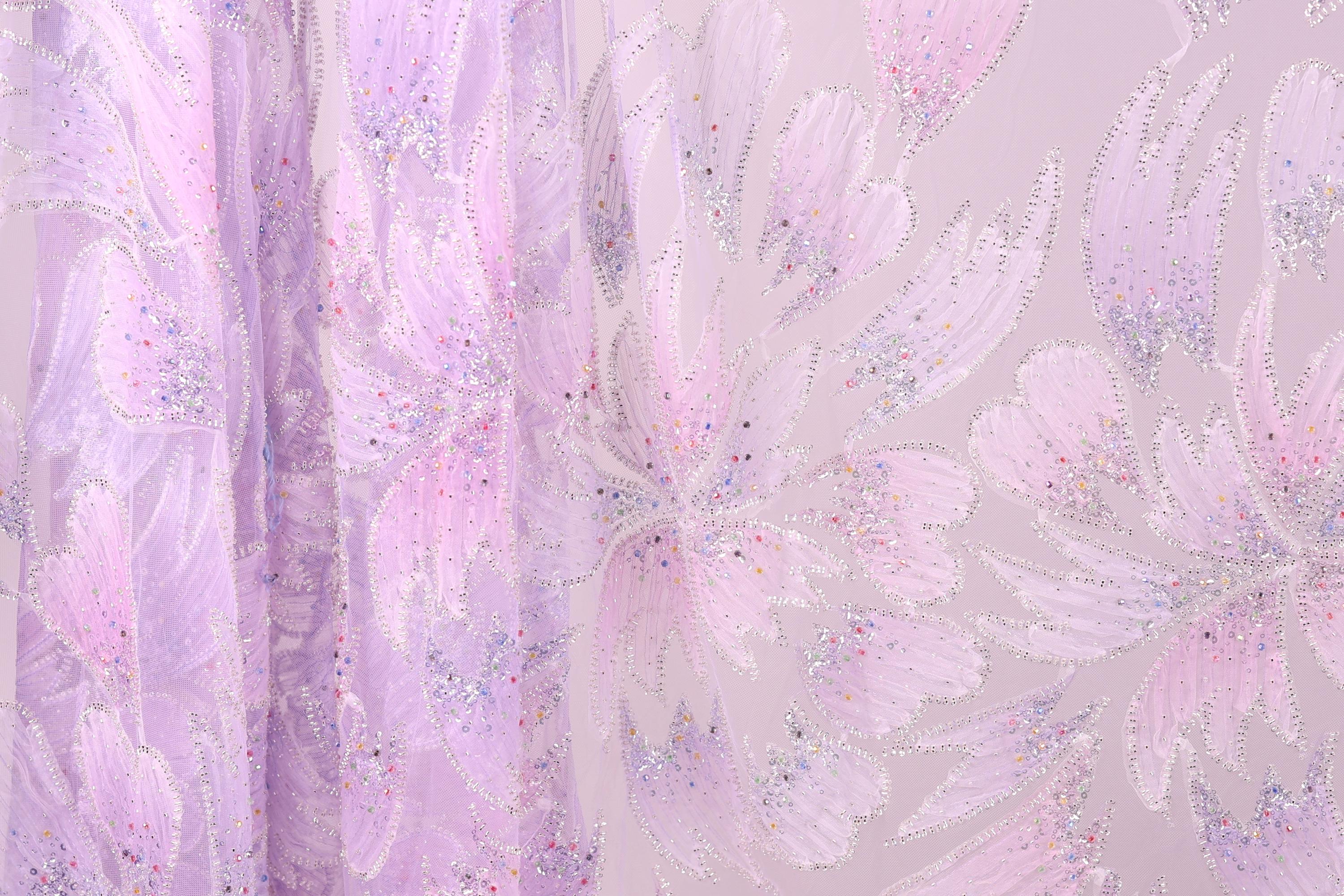 a close-up view of a fabric with a floral pattern in shades of pink and purple. The fabric appears to be made of a sheer material, with a delicate and intricate design. The pattern consists of multiple flowers and leaves, creating a visually appealing and textured appearance.