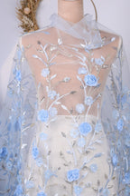 a mannequin wearing a white dress with a sheer blue floral fabric draped over it. The dress has a delicate floral pattern with blue flowers and leaves, creating a beautiful and elegant appearance.