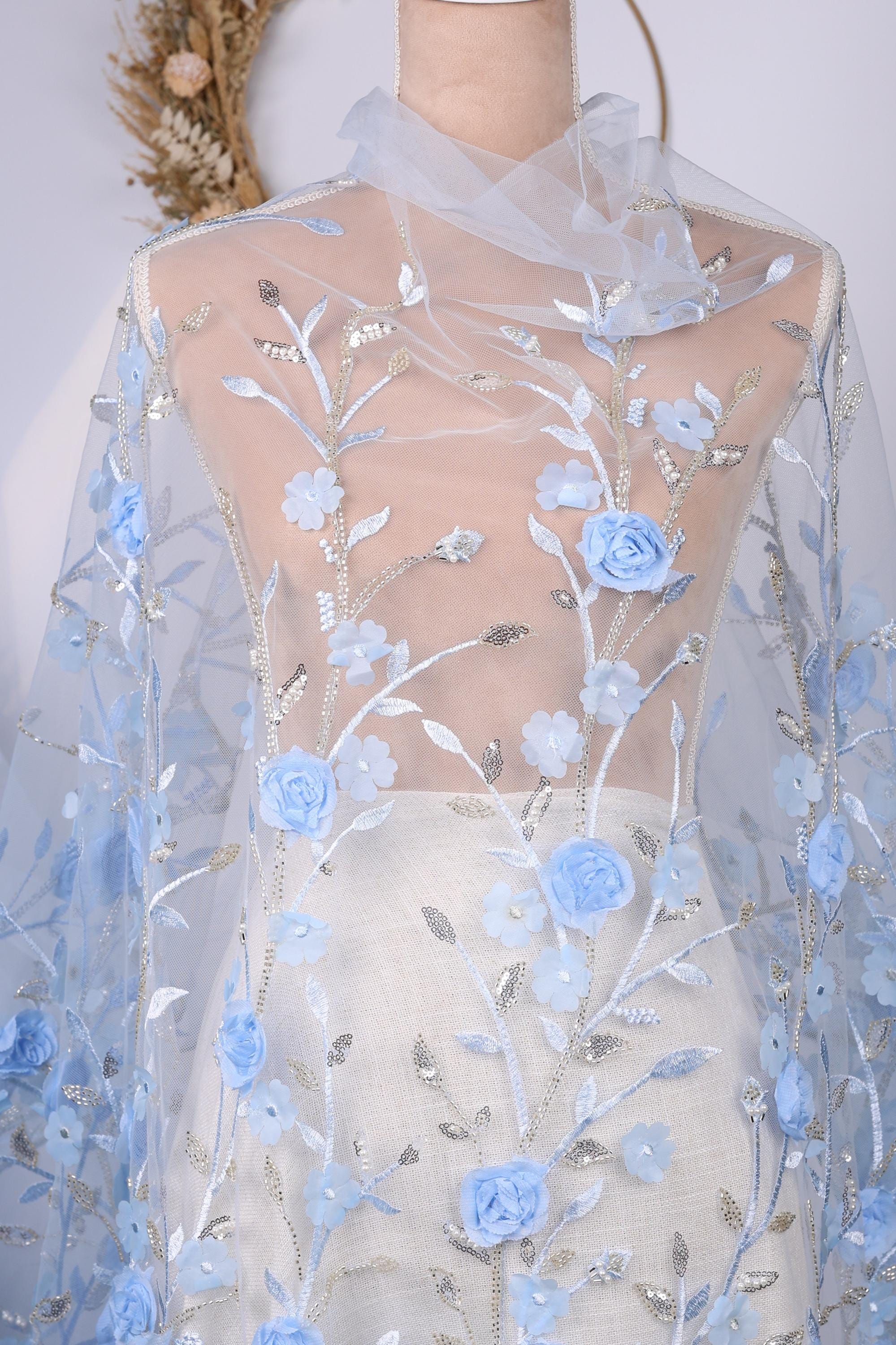 a mannequin wearing a white dress with a sheer blue floral fabric draped over it. The dress has a delicate floral pattern with blue flowers and leaves, creating a beautiful and elegant appearance.