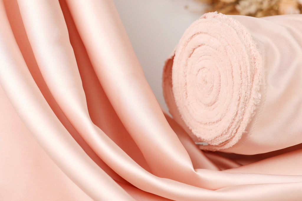 A close-up image of a soft, light pink fabric, possibly silk or satin, with a curled or rolled end visible.