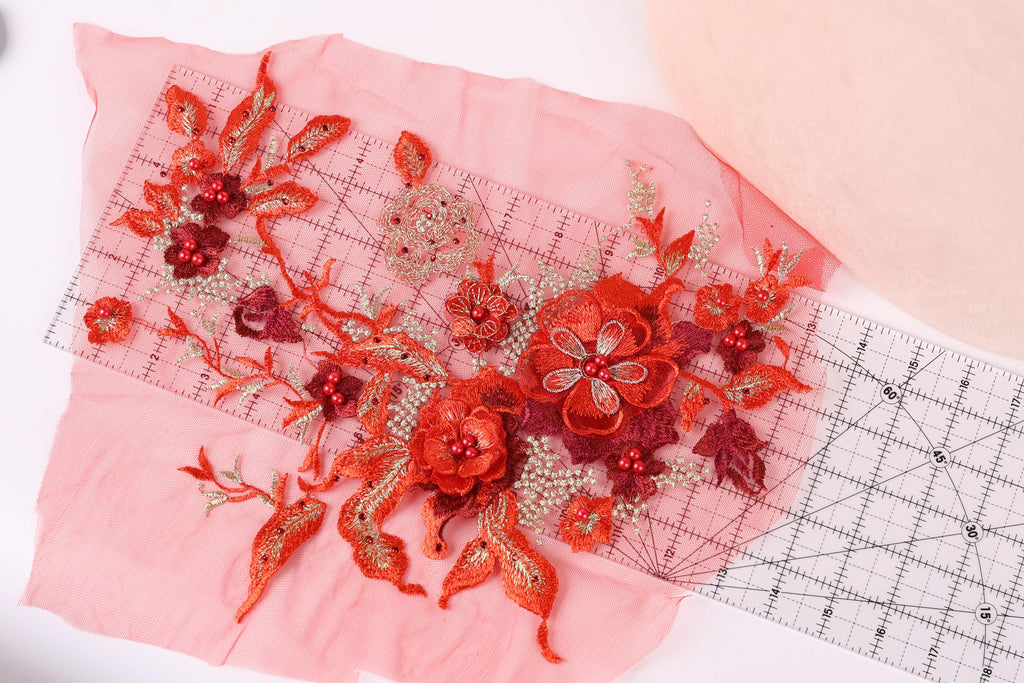 a close-up view of a pink fabric adorned with red and gold embroidered flowers and leaves. The fabric is spread out on a pink background, and a ruler is placed next to it, likely for measuring or cutting purposes.
