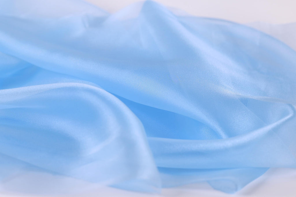The image is a close-up of a soft, light blue fabric, possibly silk or chiffon, with a smooth and flowing texture.