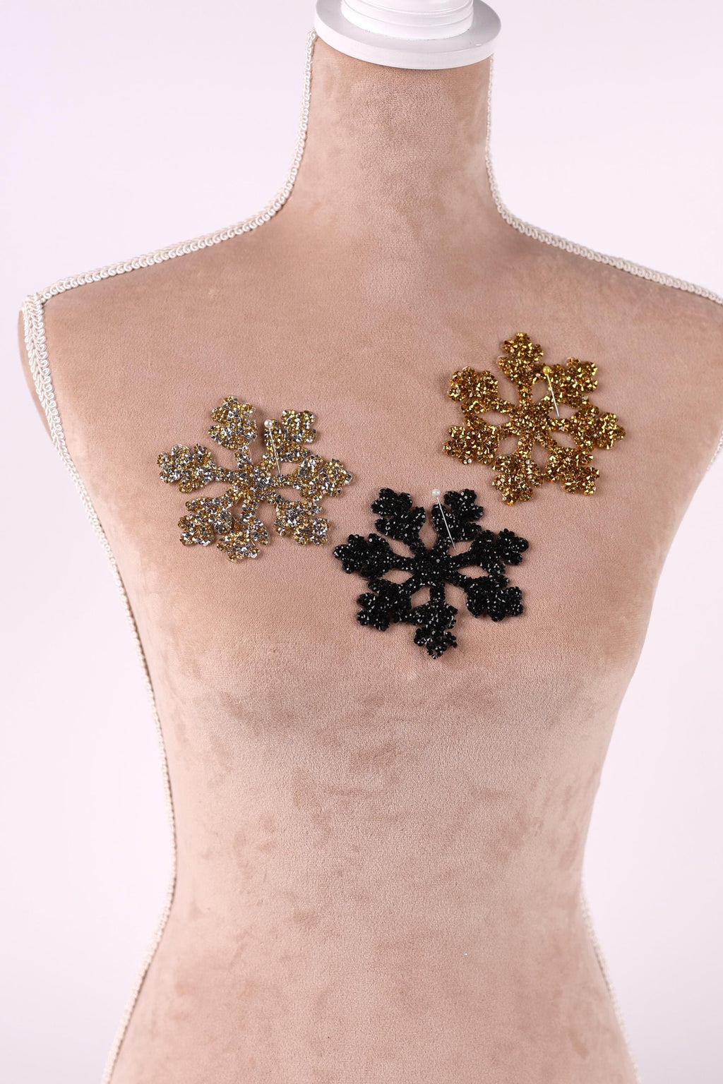 A mannequin is wearing a beige dress with three decorative snowflake-shaped embellishments in different colors - gold, black, and silver.