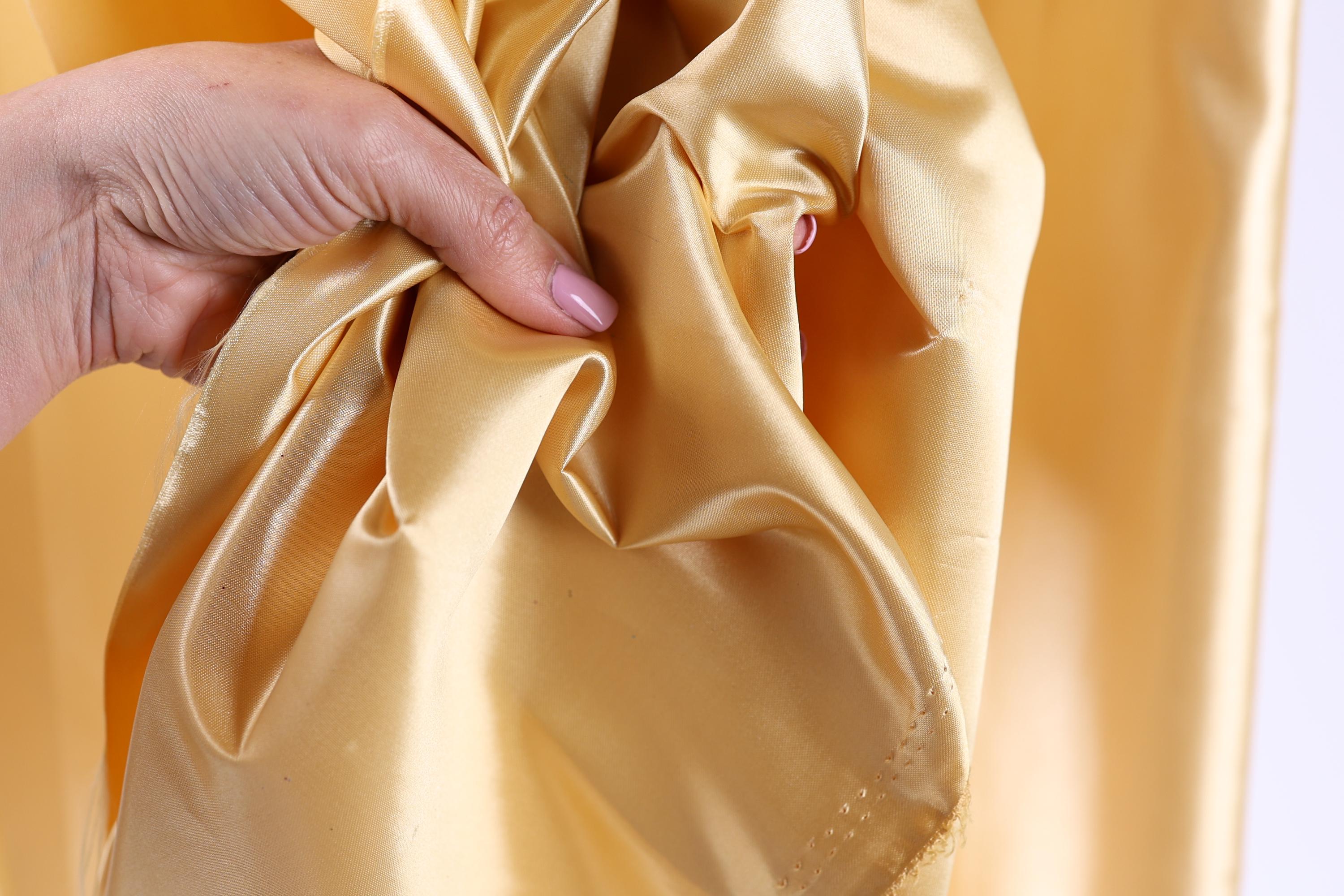 A hand is holding a piece of yellow satin fabric, with the fabric appearing to be wrinkled.