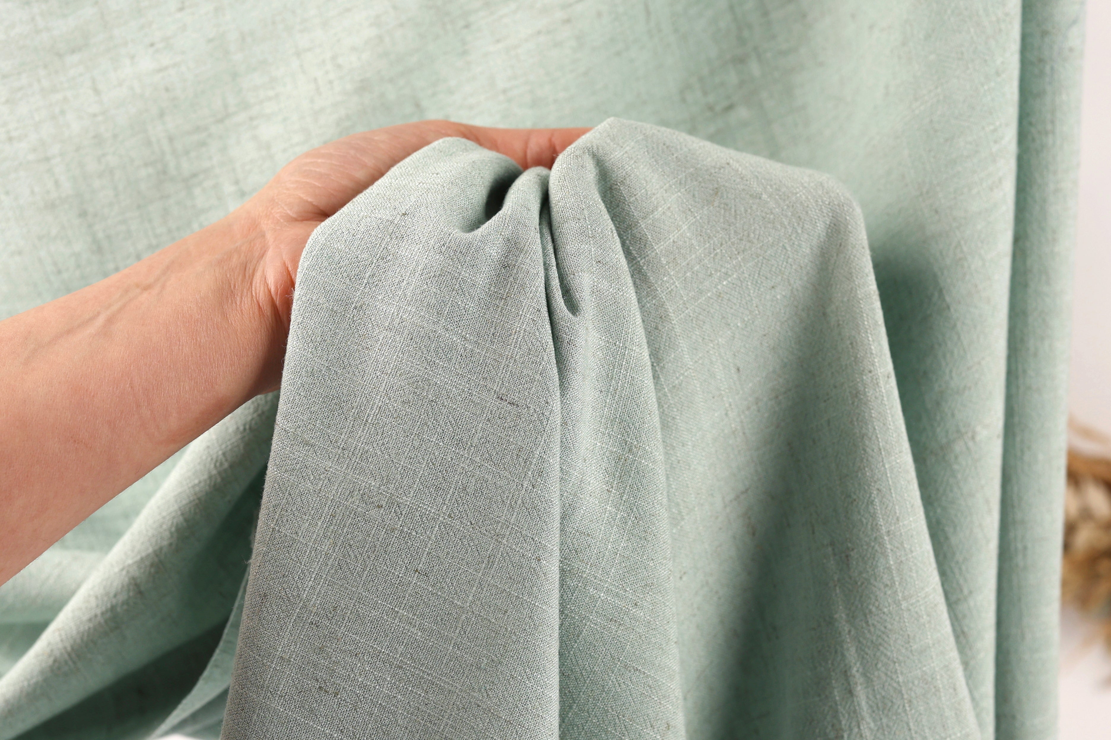 A hand is holding a light green fabric, possibly a piece of clothing or a textile, in front of a light green fabric.