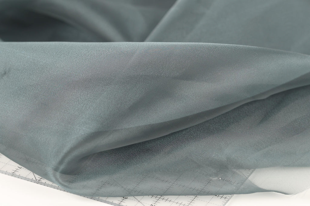 a close-up view of a piece of fabric, possibly a sheer material, with a gray color. The fabric appears to be folded or crumpled, and there is a ruler or measuring tape visible in the bottom right corner of the image.