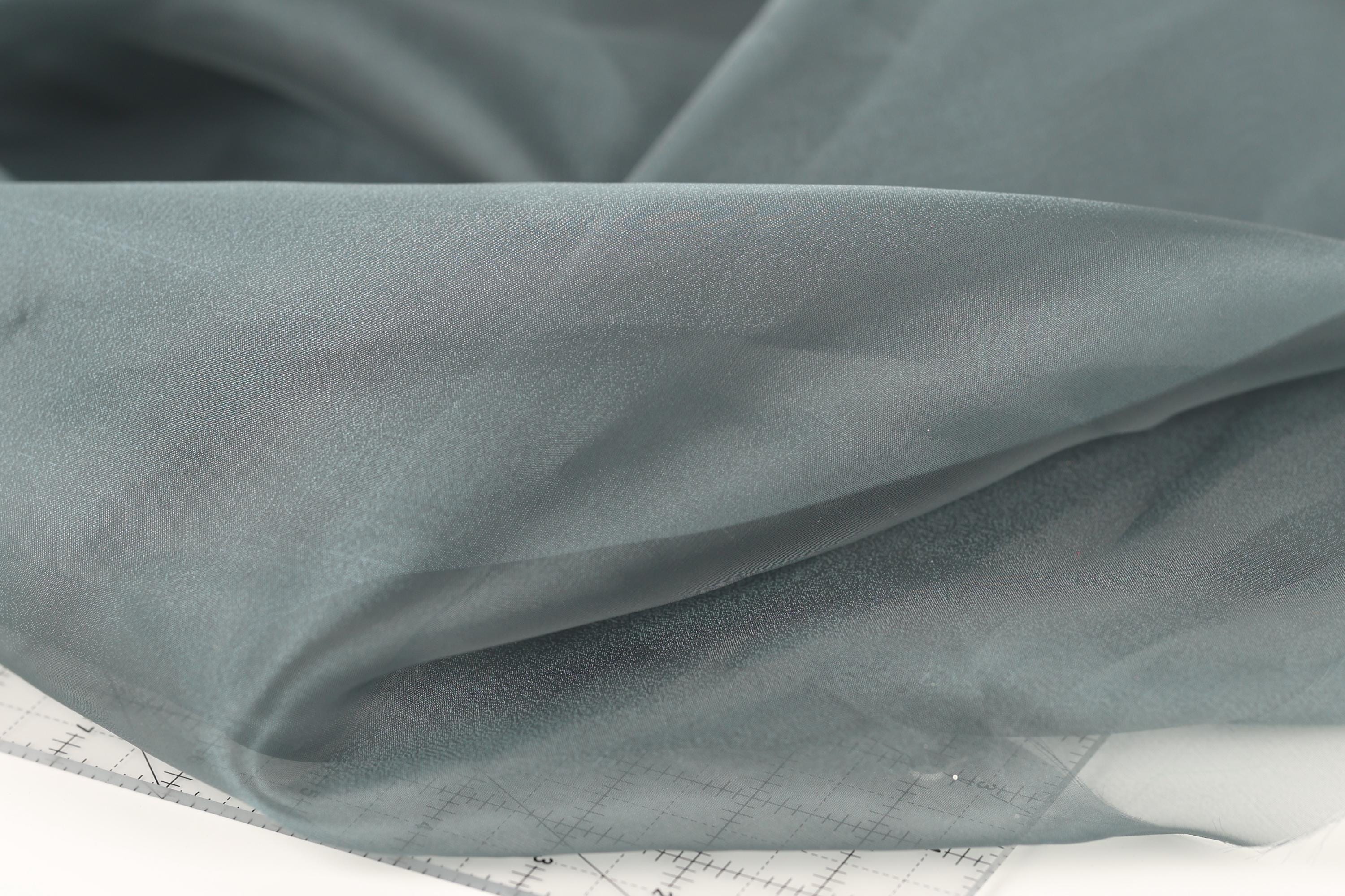 a close-up view of a piece of fabric, possibly a sheer material, with a gray color. The fabric appears to be folded or crumpled, and there is a ruler or measuring tape visible in the bottom right corner of the image.