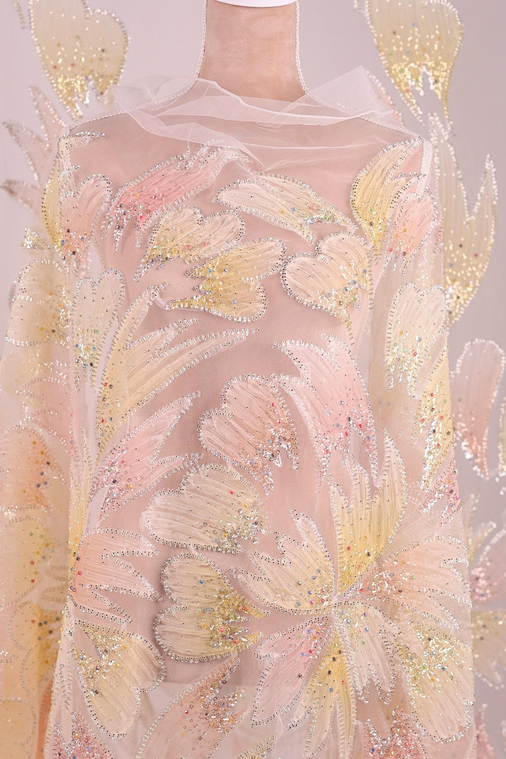 a close-up view of a delicate, intricately embroidered fabric with a floral pattern. The fabric appears to be made of sheer material, giving it a soft and delicate appearance. The embroidery is detailed and colorful, with a variety of shades of pink, yellow, and white creating a visually striking and elegant design.