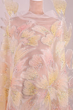a close-up view of a delicate, intricately embroidered fabric with a floral pattern. The fabric appears to be made of sheer material, giving it a soft and delicate appearance. The embroidery is detailed and colorful, with a variety of shades of pink, yellow, and white creating a visually striking and elegant design.
