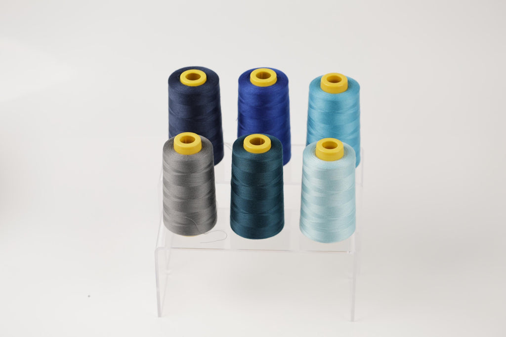 A collection of six spools of thread in various colors, including blue, gray, and light blue, arranged on a clear stand.