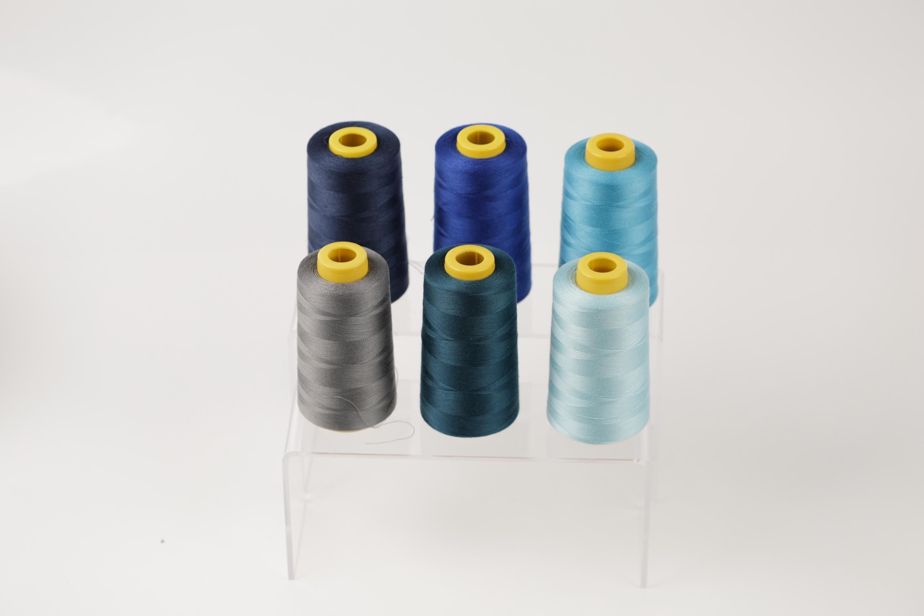 A collection of six spools of thread in various colors, including blue, gray, and light blue, arranged on a clear stand.