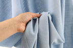 A hand is holding a piece of light blue fabric, possibly a curtain or garment, in front of a blurred background.