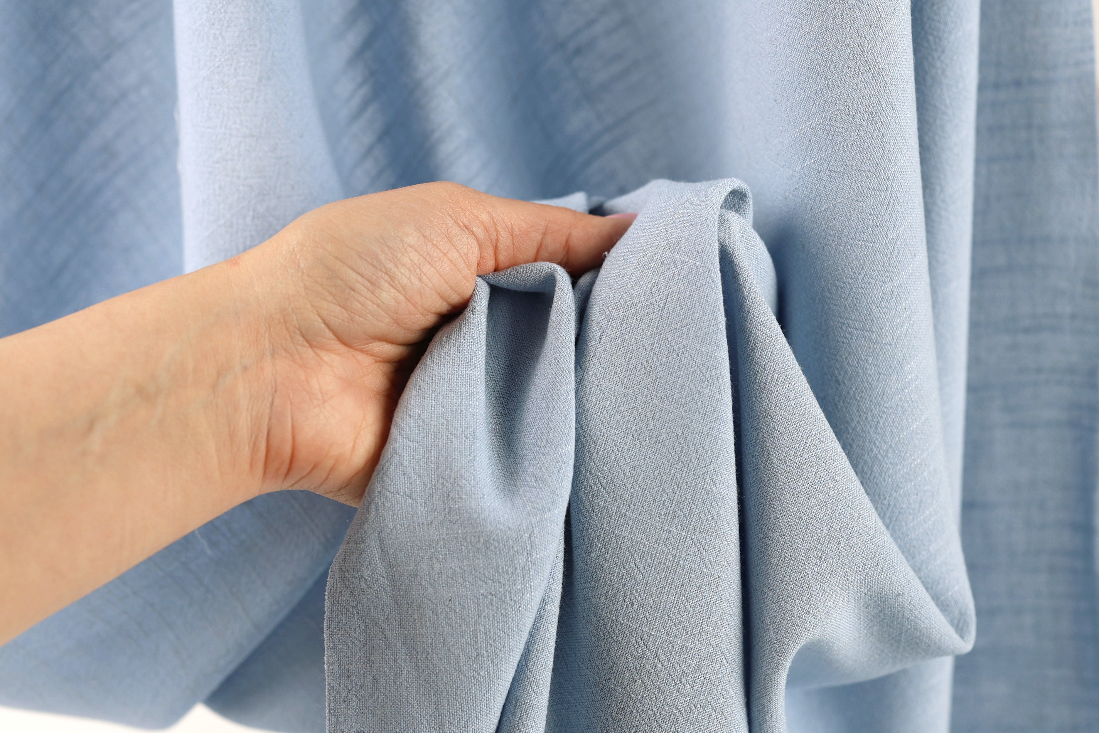 A hand is holding a piece of light blue fabric, possibly a curtain or garment, in front of a blurred background.