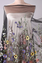 a mannequin wearing a dress with a floral pattern. The dress has a sheer overlay and is covered in a variety of colorful flowers, including yellow, purple, and pink flowers. The dress appears to be made of a sheer fabric, giving it a delicate and airy appearance.