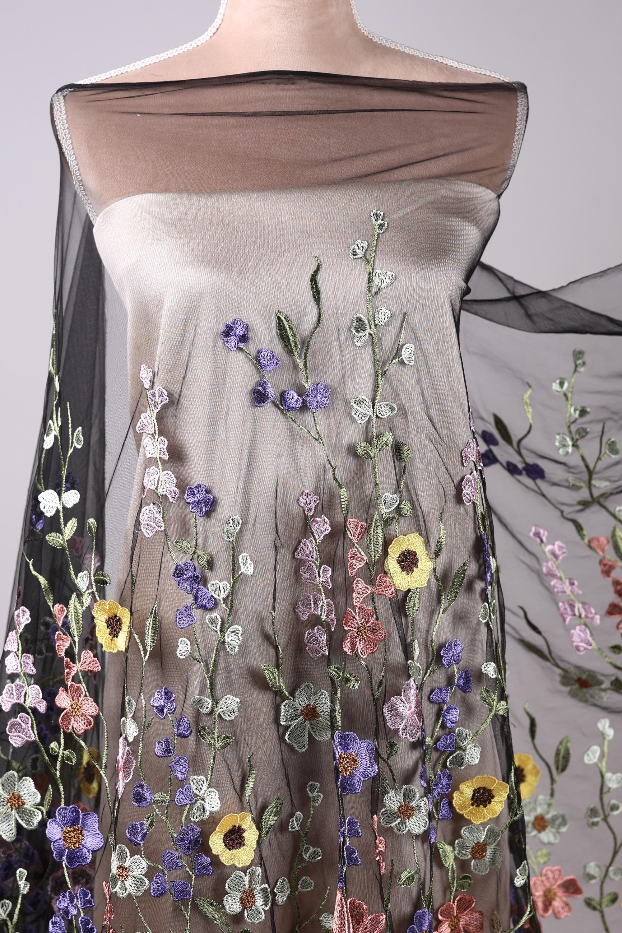 a mannequin wearing a dress with a floral pattern. The dress has a sheer overlay and is covered in a variety of colorful flowers, including yellow, purple, and pink flowers. The dress appears to be made of a sheer fabric, giving it a delicate and airy appearance.