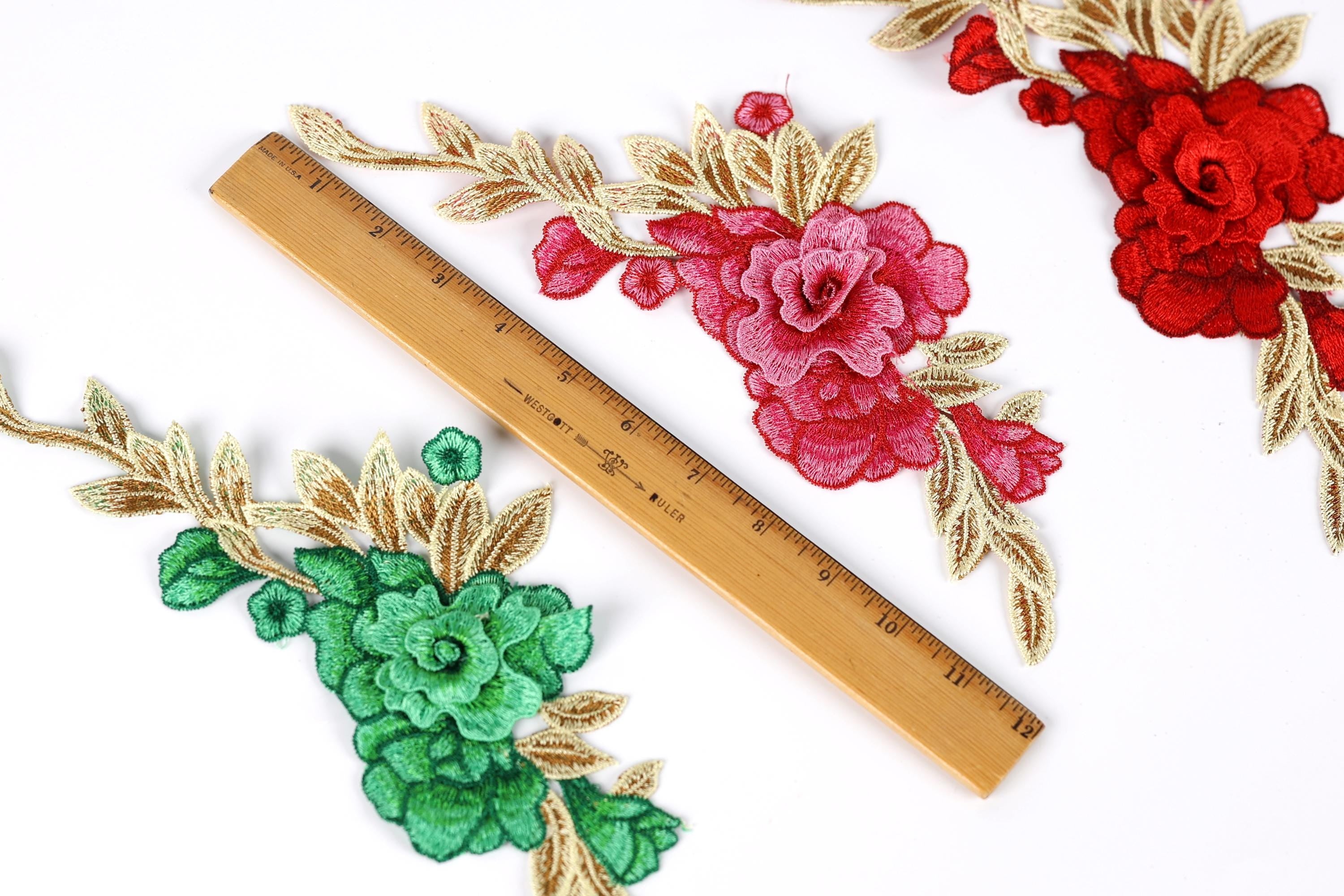 three floral embroidery pieces with different colors and designs, including red, pink, and green flowers, as well as leaves. A wooden ruler is placed between the floral pieces, indicating the size or dimensions of the embroidery.