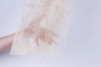 A hand is holding a piece of light-colored fabric, possibly a sheer or sheer-like material, against a plain white background.
