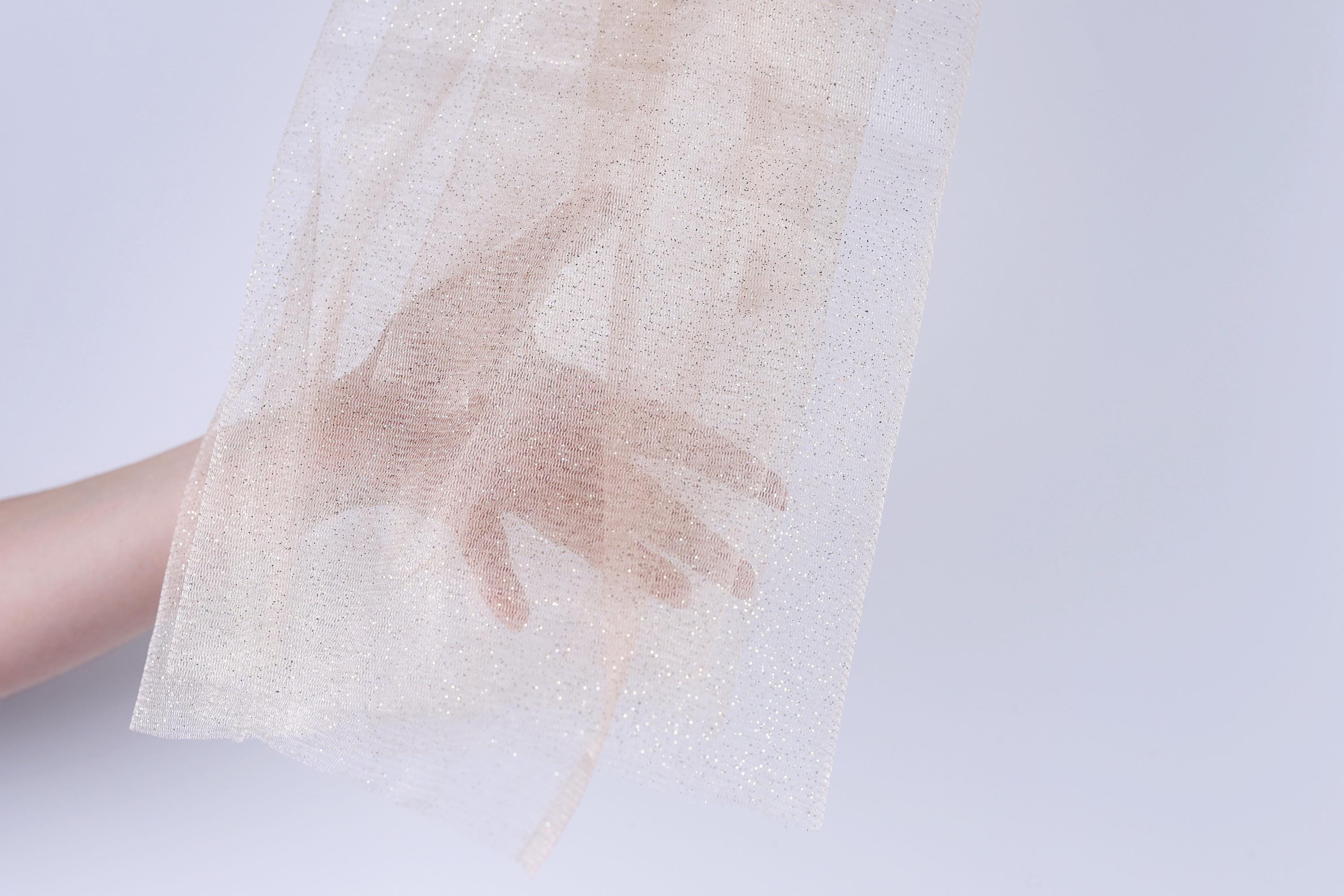 A hand is holding a piece of light-colored fabric, possibly a sheer or sheer-like material, against a plain white background.
