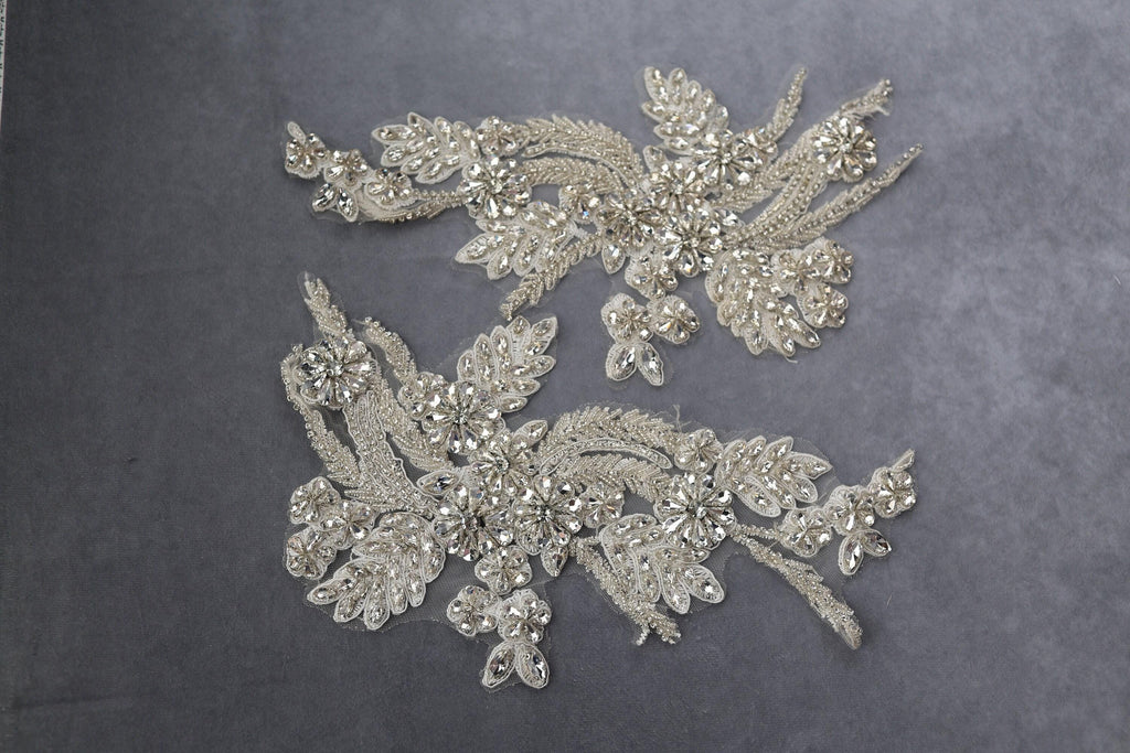 a pair of ornate, bejeweled fabric pieces with intricate designs and sparkling embellishments, including flowers and leaves, set against a gray background.