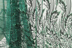 a close-up view of a fabric with a repeating pattern of green and black leaf-like designs. The pattern is intricate and detailed, with the leaves appearing to be made of a shiny material, possibly sequin or glitter. The fabric is predominantly green, with the black leaf designs providing a striking contrast. The image does not contain any text or other discernible objects, and the focus is solely on the detailed leaf pattern.