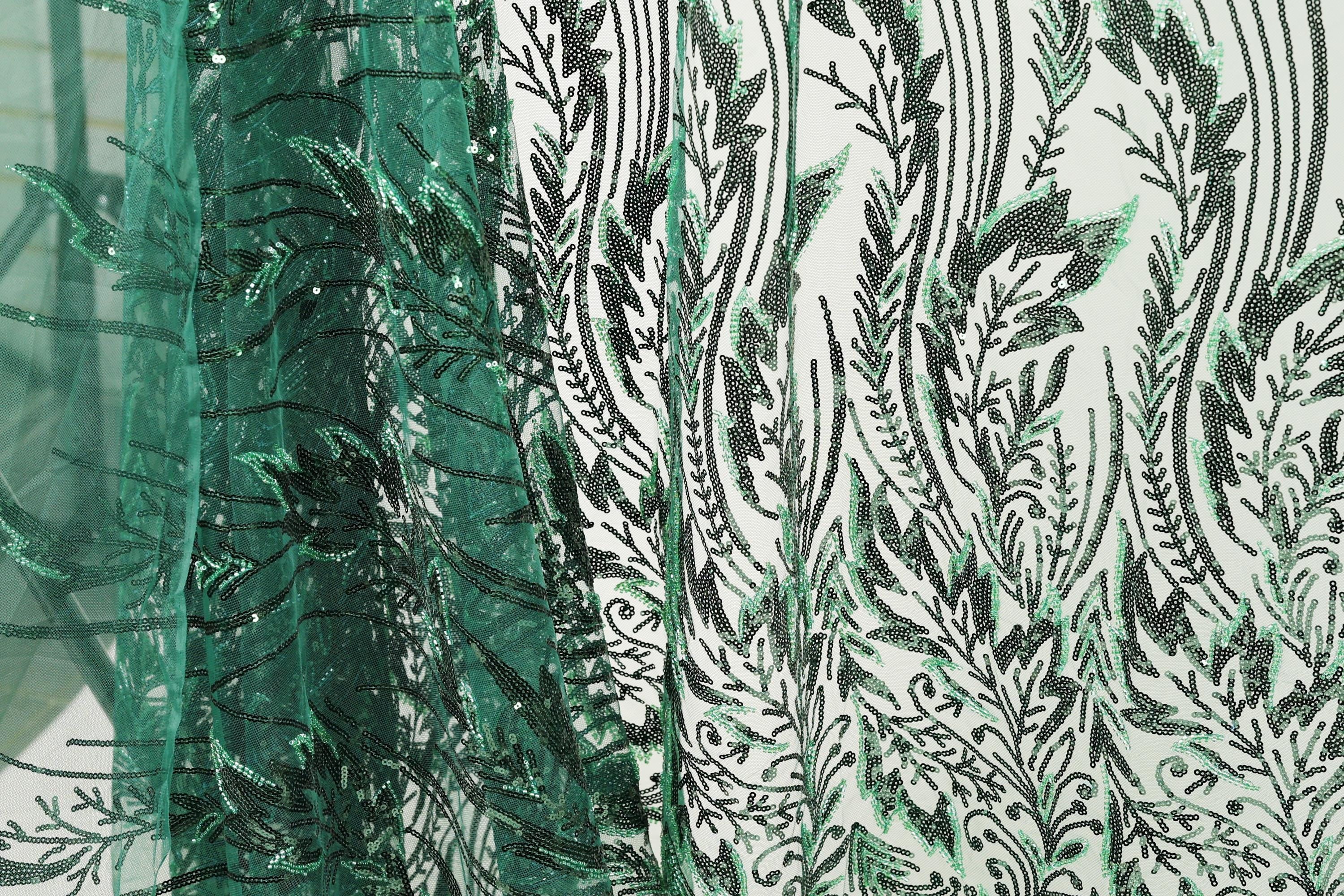 a close-up view of a fabric with a repeating pattern of green and black leaf-like designs. The pattern is intricate and detailed, with the leaves appearing to be made of a shiny material, possibly sequin or glitter. The fabric is predominantly green, with the black leaf designs providing a striking contrast. The image does not contain any text or other discernible objects, and the focus is solely on the detailed leaf pattern.