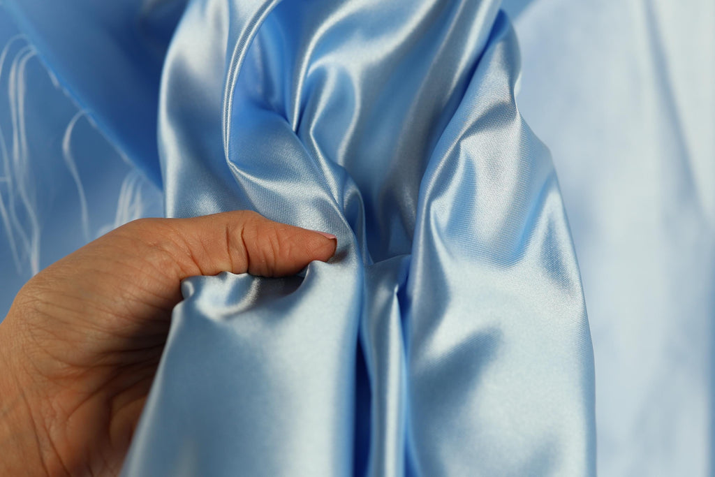 A hand is holding a piece of blue satin fabric, examining its texture and quality.