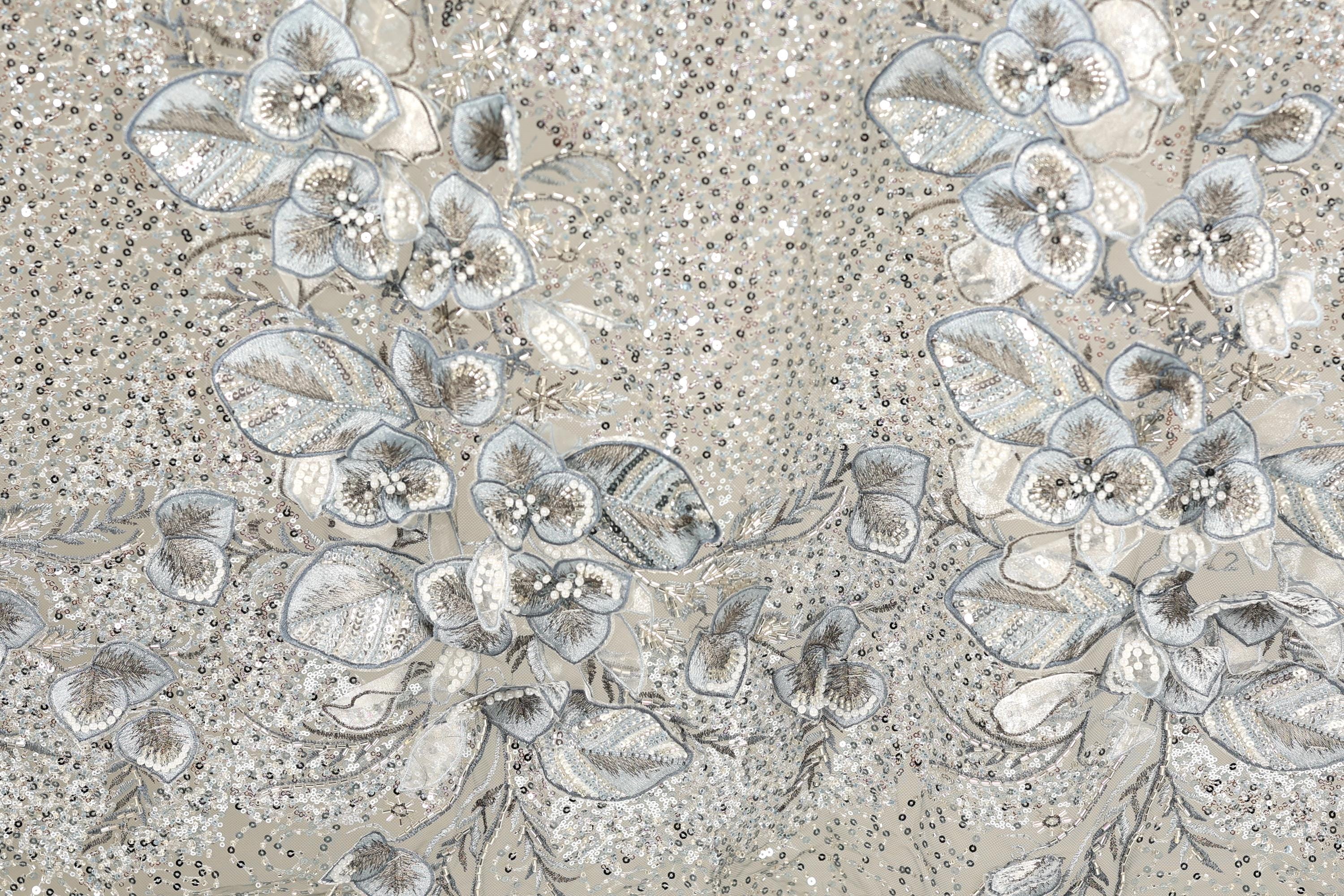 a close-up view of a fabric with a floral pattern. The fabric appears to be made of a material with a silver or metallic sheen, and it is adorned with numerous small white flowers and leaves. The pattern is intricate and detailed, showcasing the craftsmanship and artistry involved in creating the fabric.