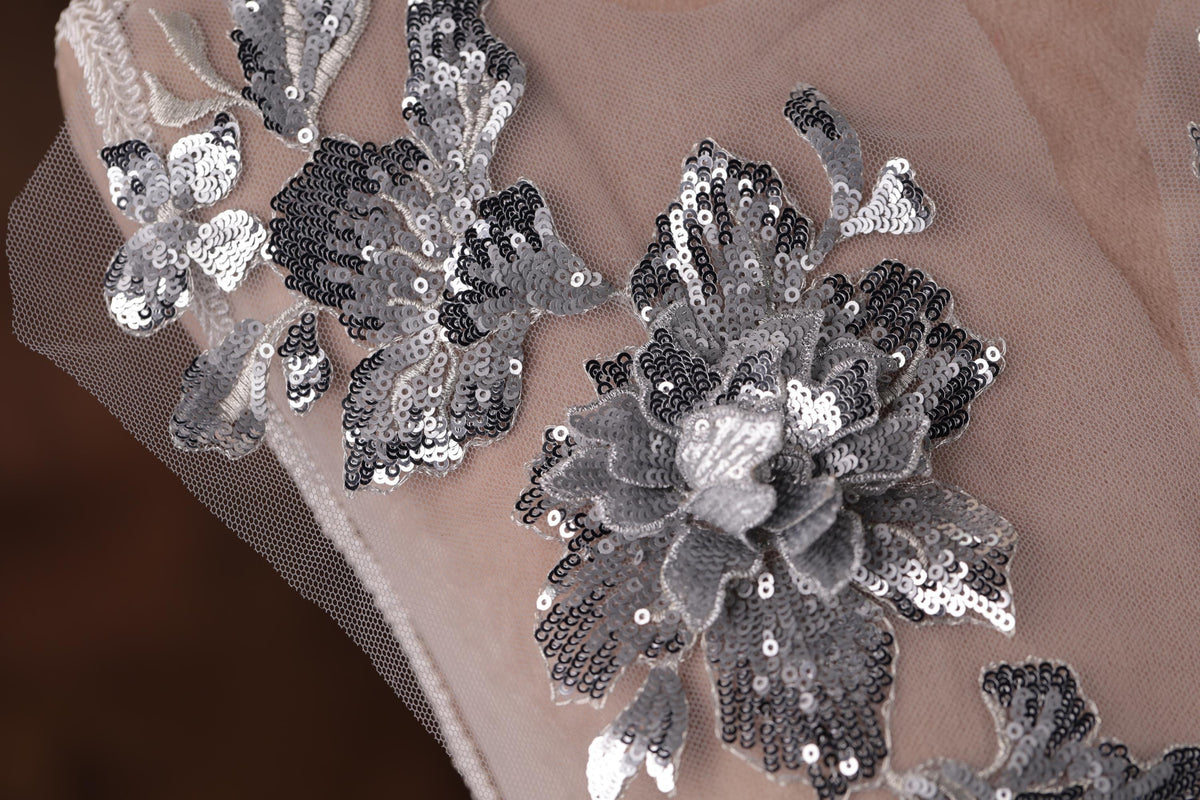 a close-up view of a delicate, intricate piece of lace or fabric adorned with a floral pattern. The pattern consists of small, black and silver sequins that create a visually striking and elegant design. The lace appears to be a part of a larger piece of clothing or textile, with the floral pattern adding a touch of sophistication and detail to the overall design.
