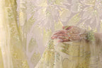 A close-up view of a hand holding a piece of fabric with a floral pattern, against a background of a white fabric with green and yellow floral designs.