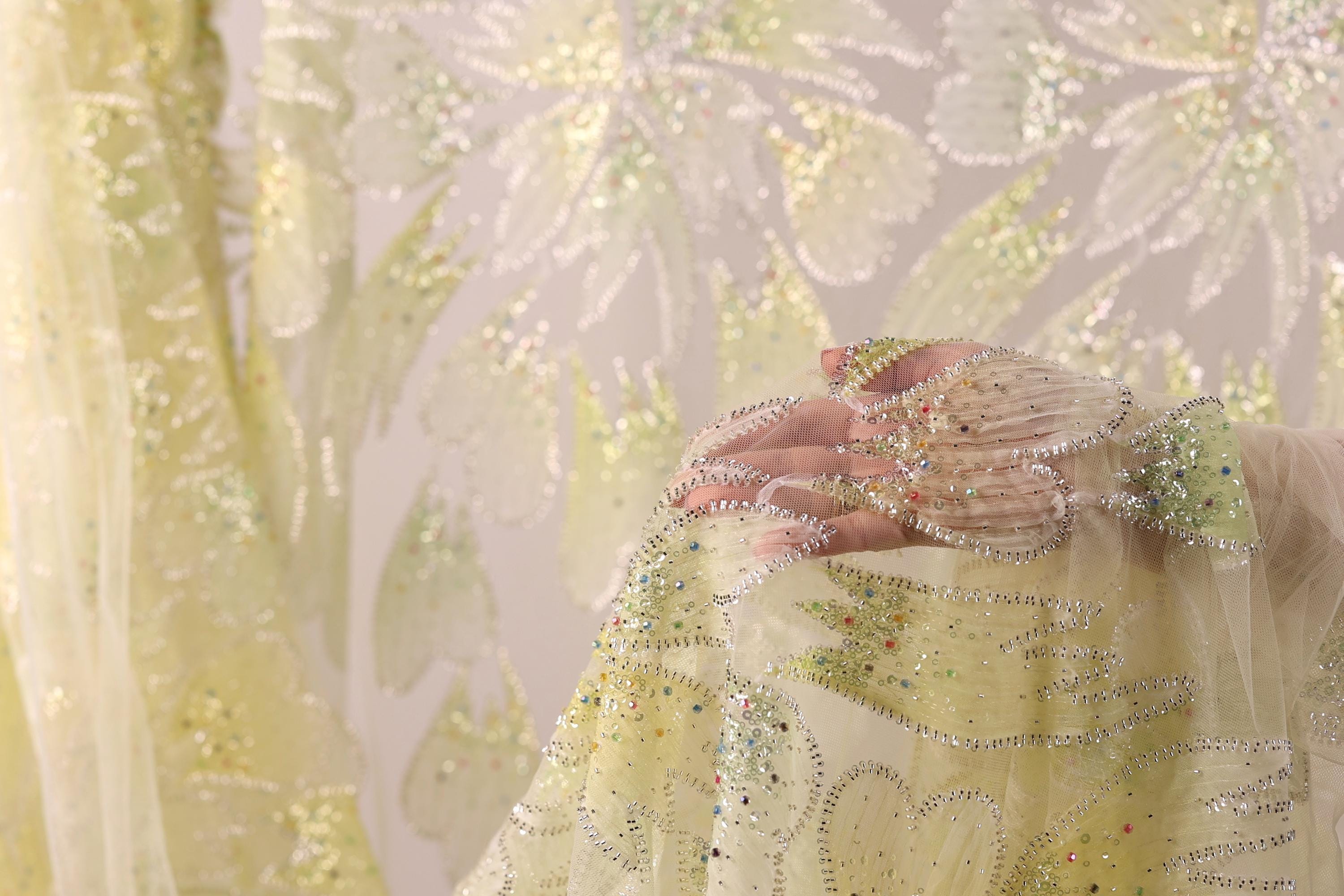 A close-up view of a hand holding a piece of fabric with a floral pattern, against a background of a white fabric with green and yellow floral designs.