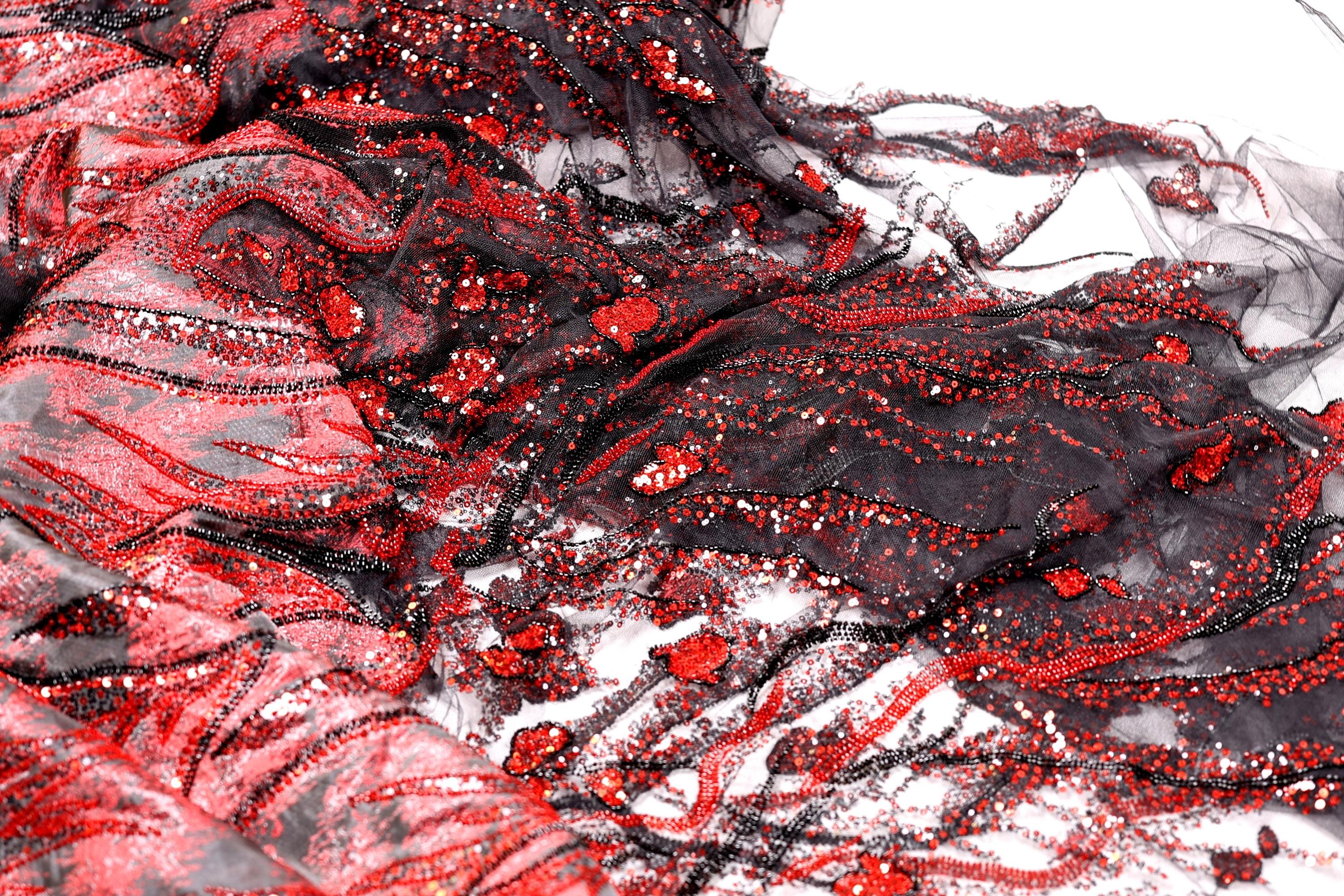 The image is a close-up of a fabric with a striking pattern of red and black sequins and lace. The fabric appears to be a sheer material, with a mix of black and red colors. The pattern is intricate and detailed, with the sequins and lace adding texture and depth to the fabric. The image does not contain any discernible objects or actions, and the focus is solely on the fabric and its design.