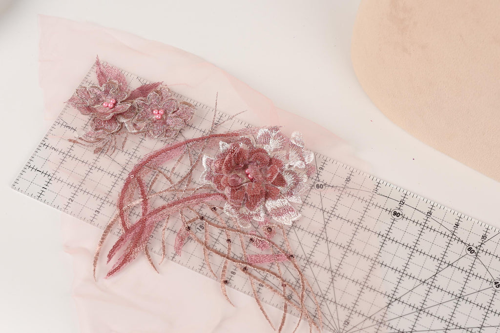 a close-up view of a pink and white floral design, including two large pink flowers with white centers and delicate petals, as well as smaller pink flowers with white centers. The design is displayed on a transparent ruler, which is placed on a white surface.
