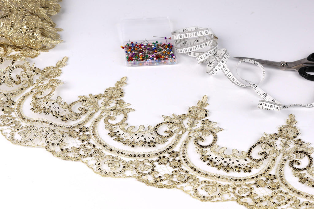 a close-up view of a decorative gold lace trim, with a spool of thread, a box of sewing pins, and a pair of scissors on a white background.
