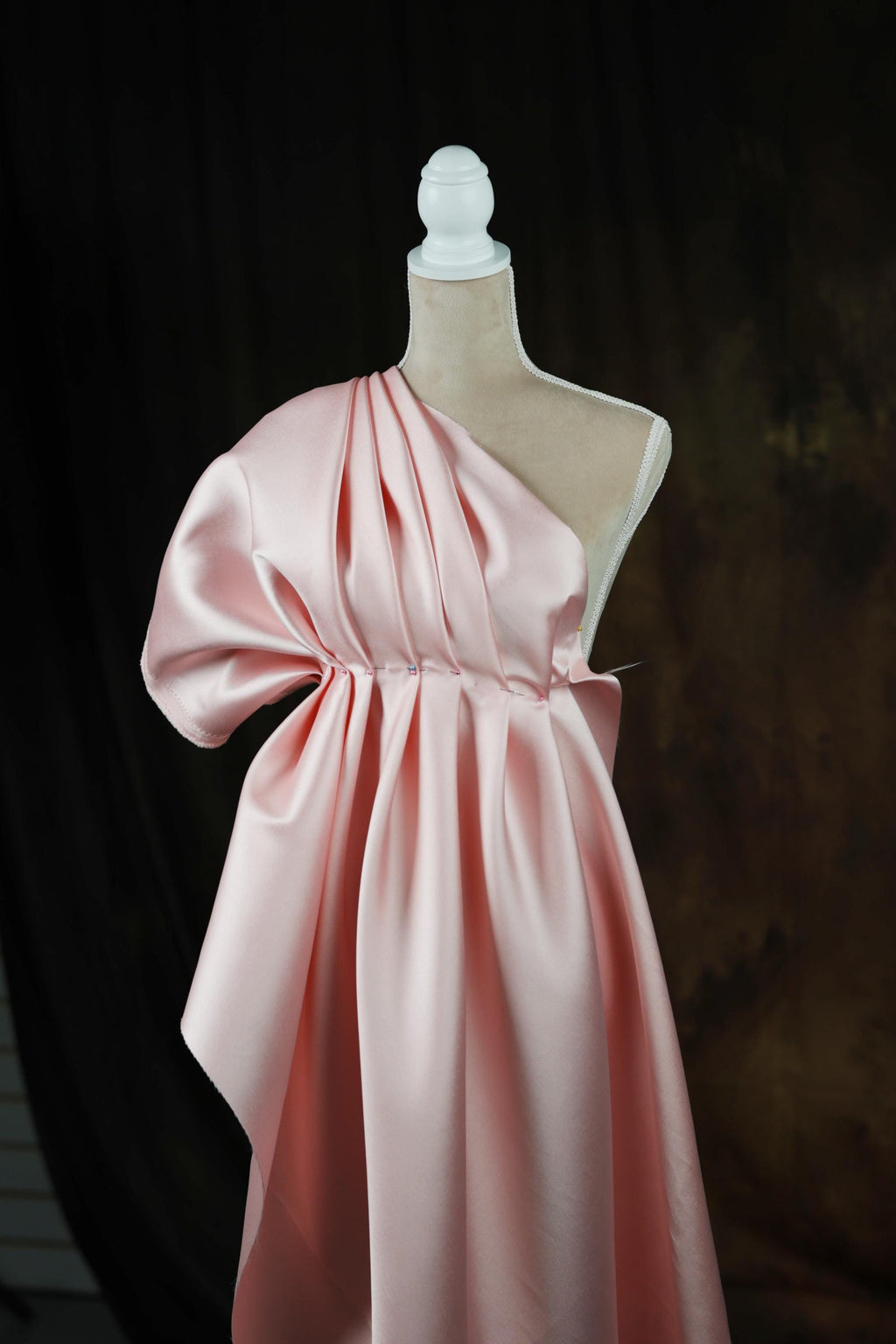 A pink satin dress with a draped, flowing silhouette is displayed on a mannequin head against a dark background.