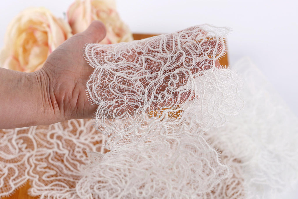 A hand is holding a piece of white lace with a floral pattern, against a blurred background of more lace pieces.